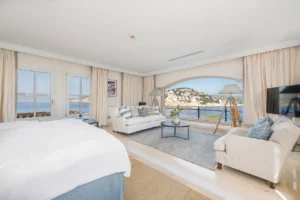 Blick übers Bett der Mastersuite in den Wohnbereich mit viel Tageslicht und Meerblick.